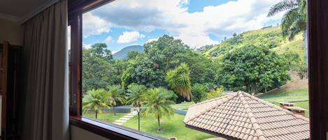 A window view of a lush garden with palm trees, a picnic table, and a tiled roof structure.