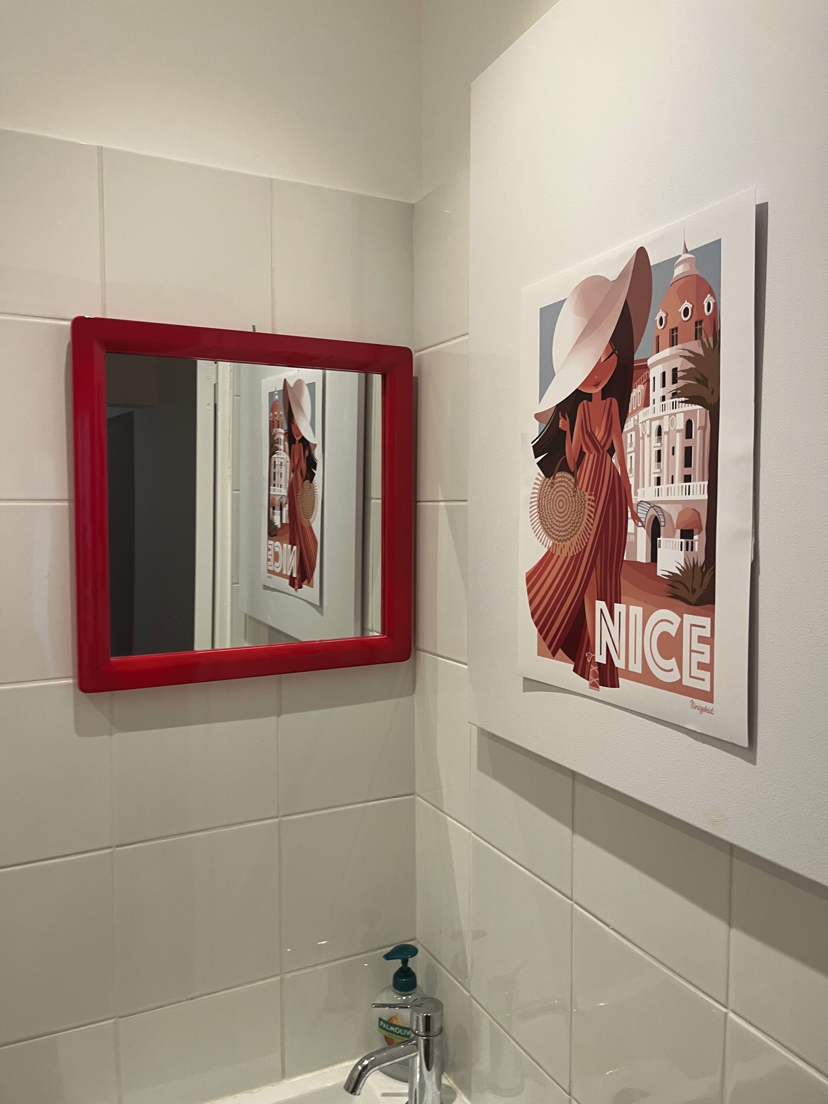 A bathroom with white tiles, a red-framed mirror, a sink with a soap dispenser, and a poster on the wall.