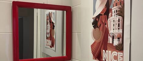 A bathroom with white tiles, a red-framed mirror, a sink with a soap dispenser, and a poster on the wall.