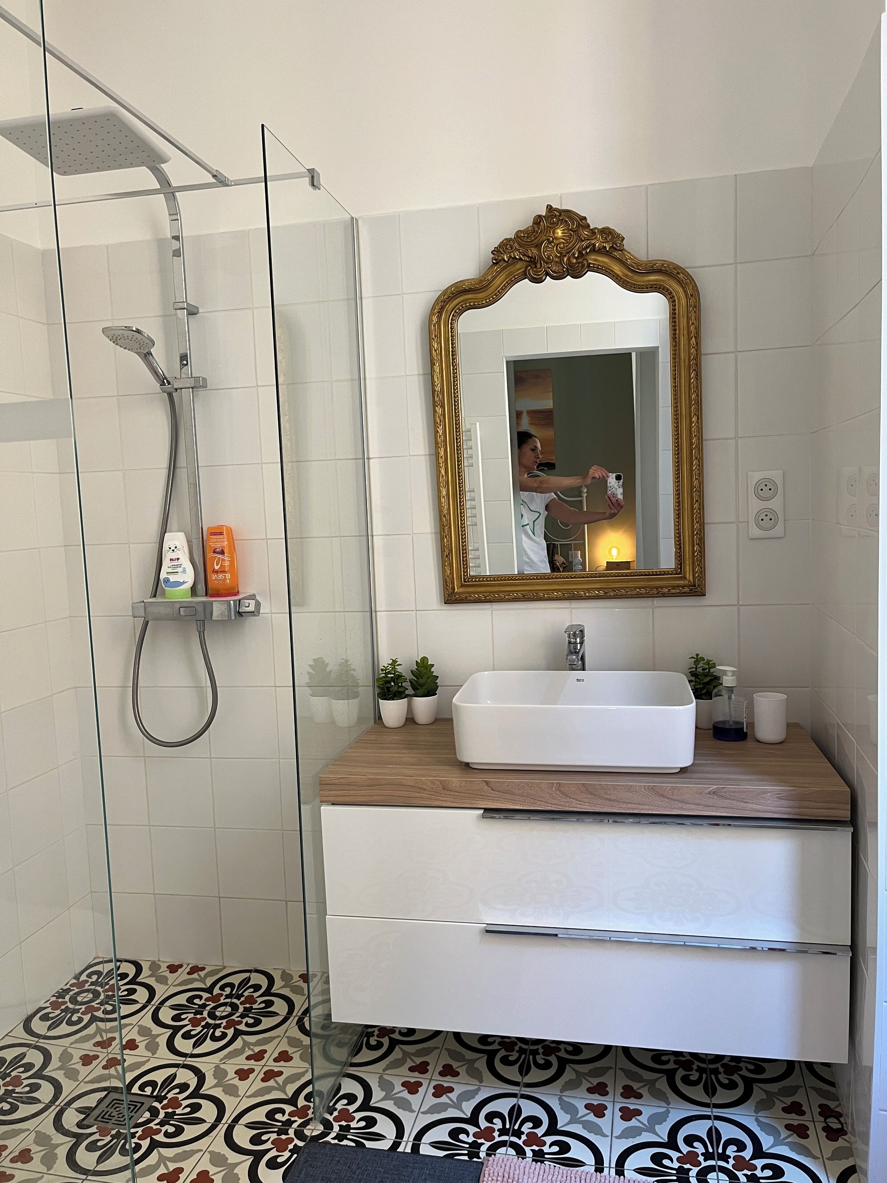 Un baño con cabina de ducha de vidrio, un lavamanos blanco con encimera de madera, un espejo con marco dorado y baldosas de suelo con diseño.