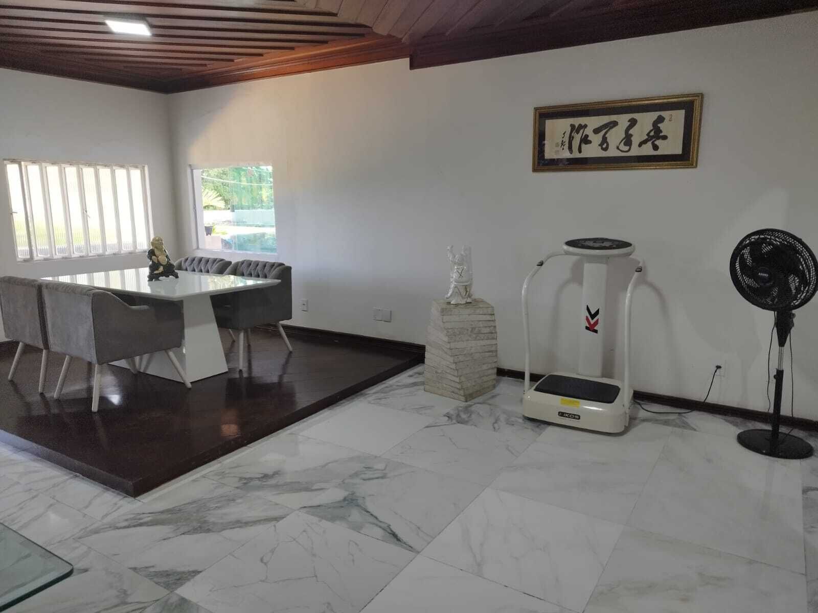 Un comedor moderno con suelo de mármol, una mesa blanca y sillas grises. Se observa una estatua sobre un pedestal, un dispensador de agua y un ventilador de pie.