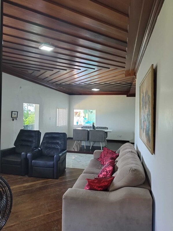 A room with a wooden ceiling, a beige sofa with a red pillow, two black leather chairs, a dining table with chairs, and a window with a view of greenery. - Casa com Piscina 50000 lts Área Gourmet (Salinópolis)