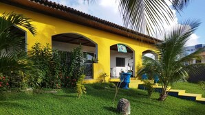 Un bâtiment jaune avec un toit en tuiles, un jardin avec de l’herbe verte et des plantes, et un drapeau bleu.