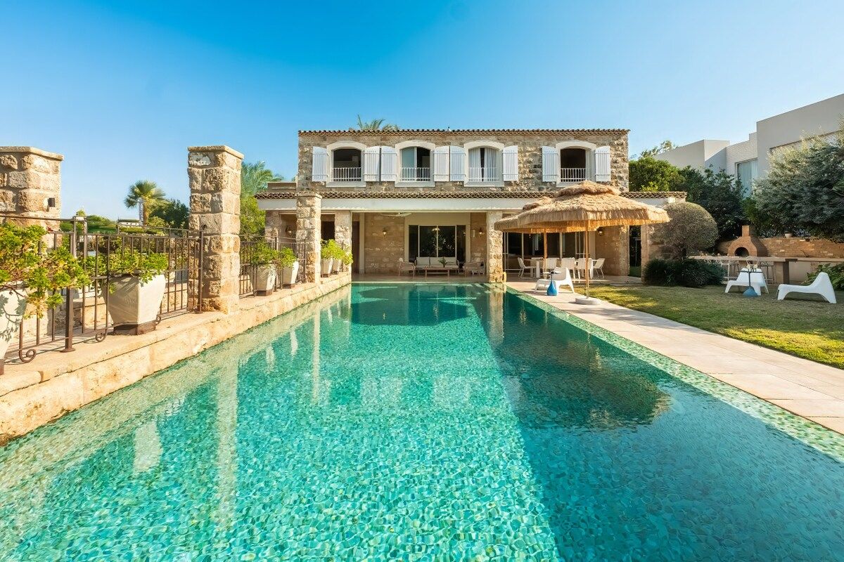 A pool with a clear blue water surface, a stone building with a balcony, and a thatched-roof structure.