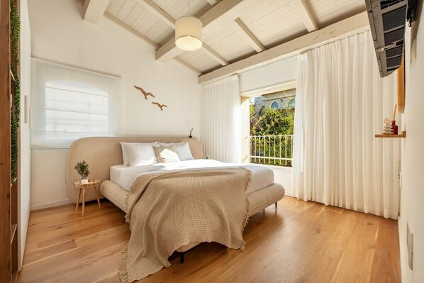 A bedroom with a bed, a small bedside table, a window with curtains, and a view of greenery outside.