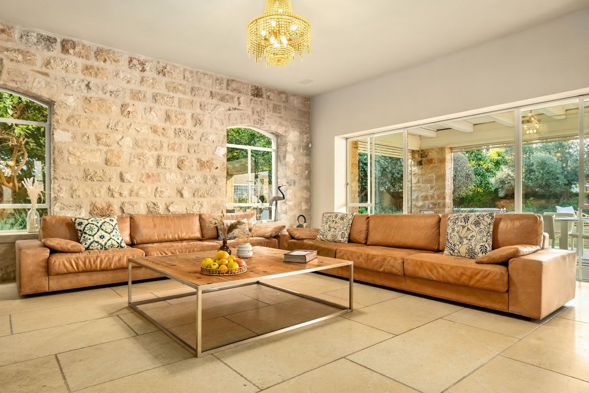 A spacious living room with a stone wall, leather sofas, a glass coffee table, and a chandelier.