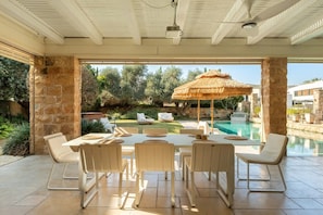 An outdoor dining area with a pool view, stone pillars, and a thatched umbrella.