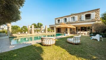 Un espace aménagé au bord de la piscine, avec un bâtiment en pierre, du mobilier d’extérieur et un ciel dégagé.