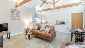 A cozy living room with a brown leather sofa, a TV, and a small table with a lamp. A kitchen with white cabinets and a wooden ceiling is visible in the background. - The Stables - UK46003 (Cranswick, near Driffield)