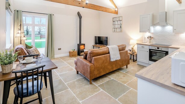 A spacious living area with a fireplace, a sofa, a dining table with chairs, and a kitchen with white cabinets and a stove. - The Stables - UK46003 (Cranswick, near Driffield)