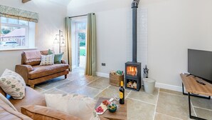 A cozy living room with a stone floor, a brown leather sofa, a wooden coffee table with snacks, a TV, and a wood-burning stove. - The Stables - UK46003 (Cranswick, near Driffield)