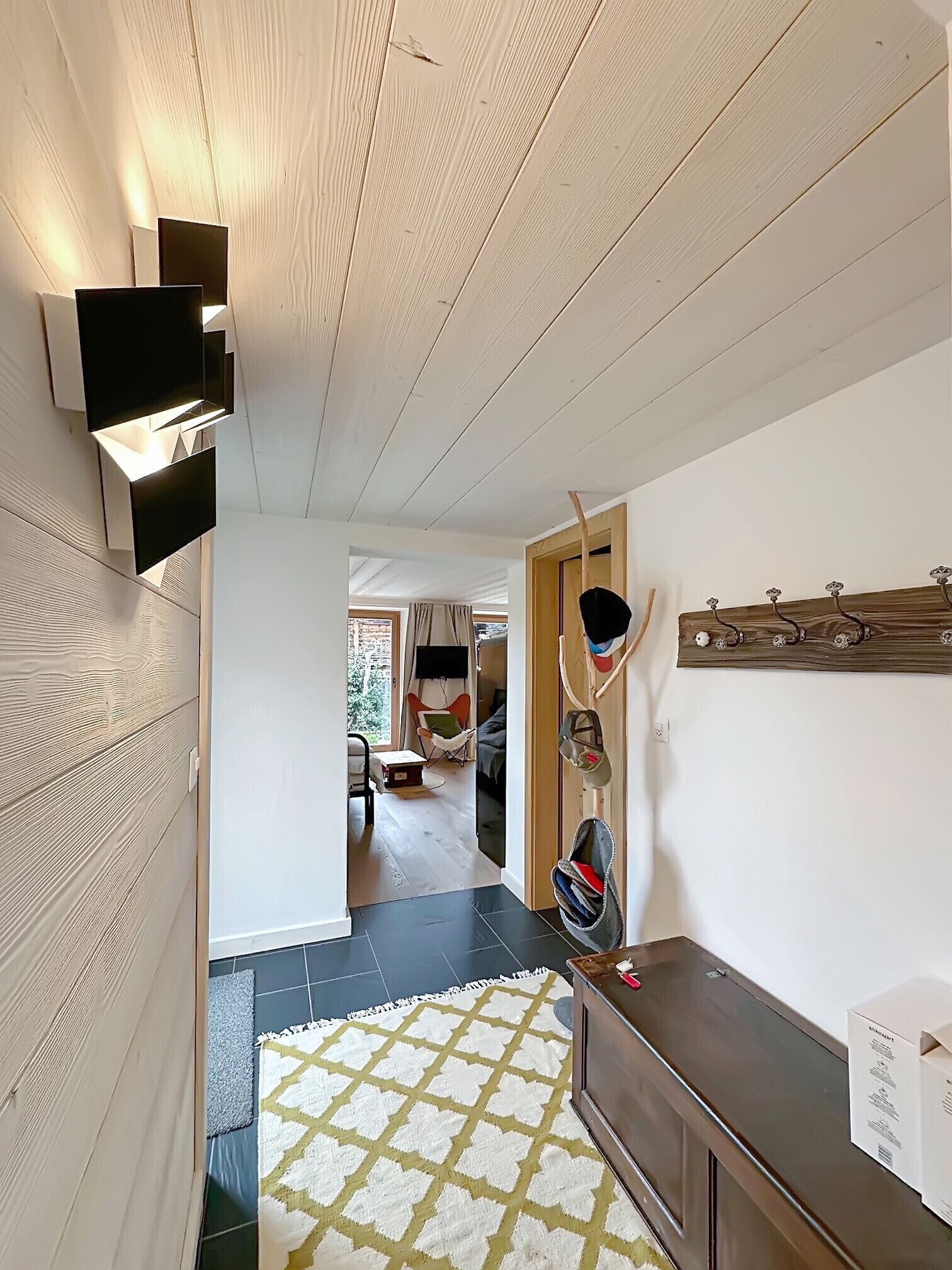 A hallway with wooden paneling, a patterned rug, a coat rack, and a glimpse of a living area with a chair and a table.