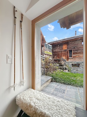 A room with a bench, wooden skis, a stone wall, and a view of a rustic building.