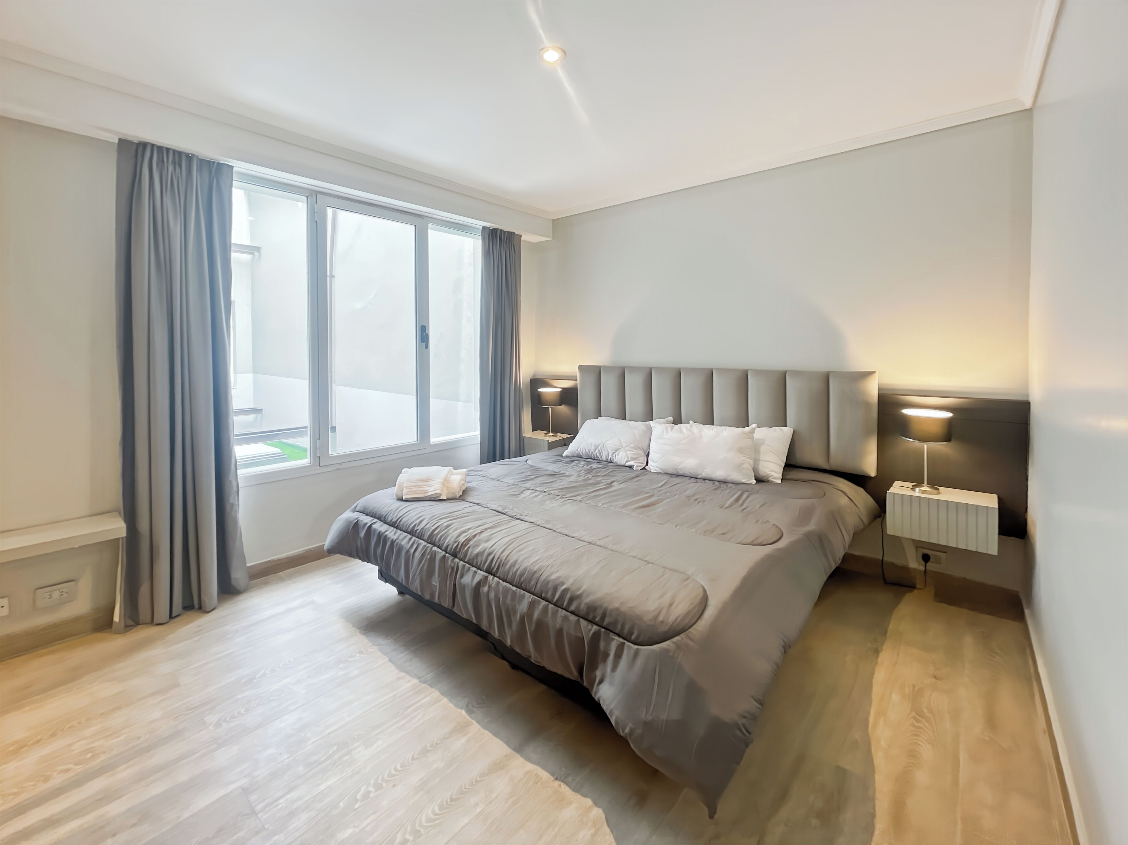 Une chambre moderne avec un grand lit, deux lampes de chevet et une vue sur un espace extérieur verdoyant par la fenêtre.