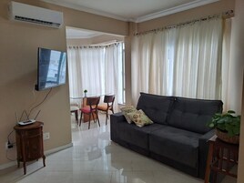 A living room with a sofa, a TV mounted on the wall, a dining table with chairs, and a plant.
