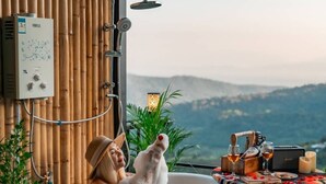 A bathtub filled with bubbles, a water heater, and a table with drinks and snacks on a balcony with a view. - Mankhao Homestay (Mae Rim)