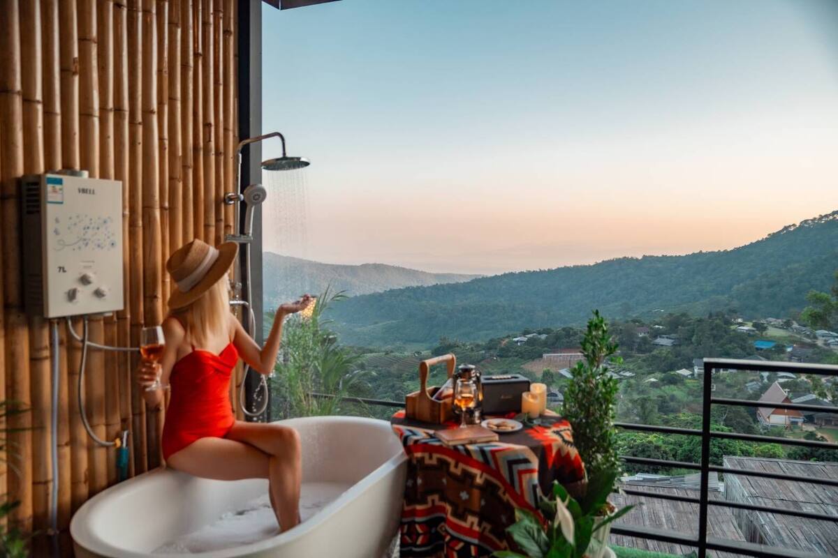 A woman in a red dress sitting in a bathtub with a showerhead above, overlooking a mountainous landscape.