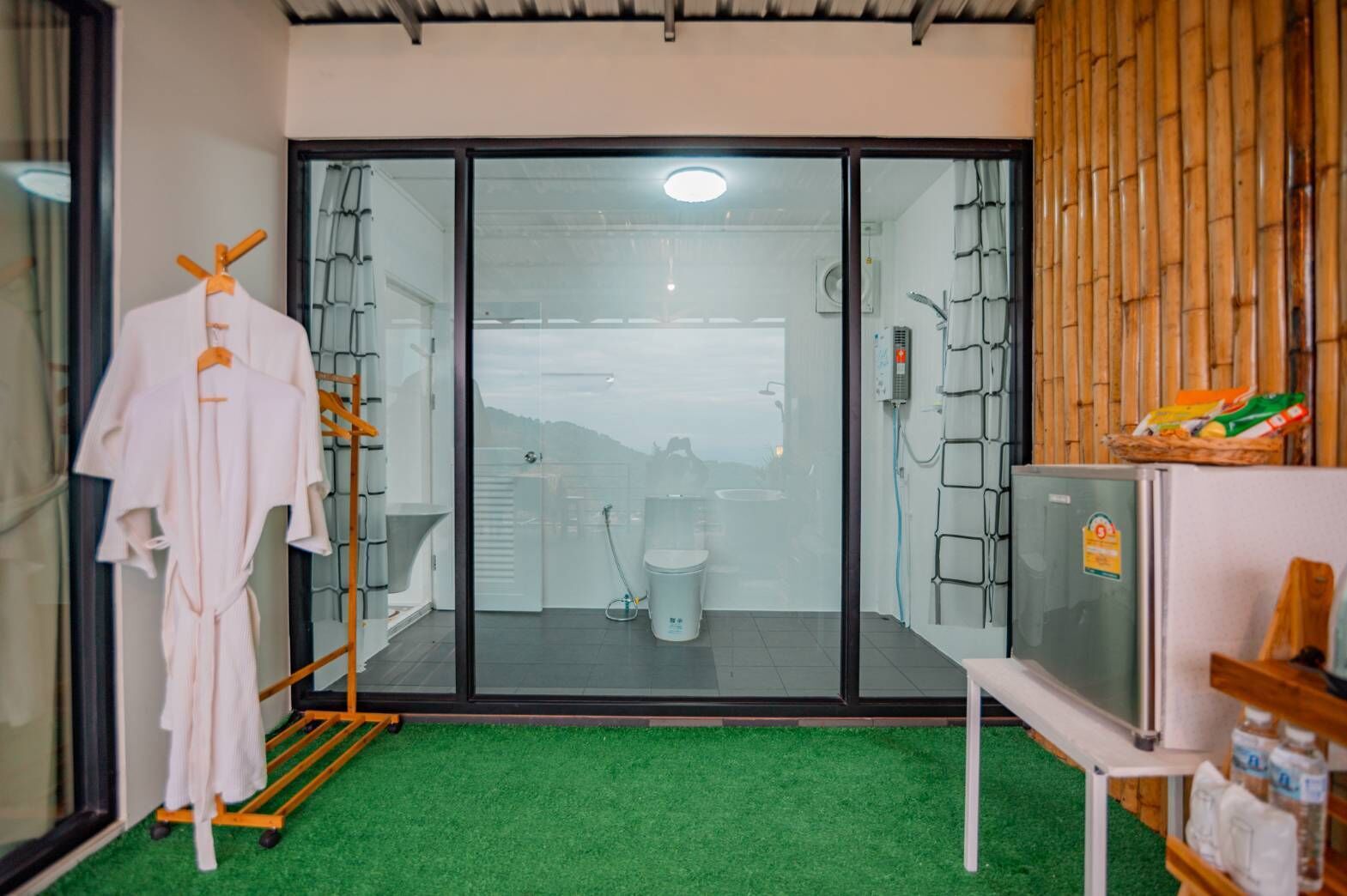 A bathroom with a glass door, a shower, and a toilet. There is a rack with towels and a television on a stand.