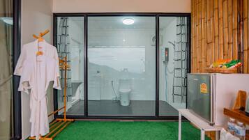 A bathroom with a glass door, a shower, and a toilet. There is a rack with towels and a television on a stand.