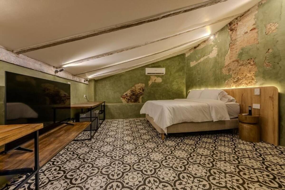 Una habitación pequeña y acogedora con una cama, un escritorio y una silla. Las paredes tienen una textura interesante y se observa una rejilla de ventilación.