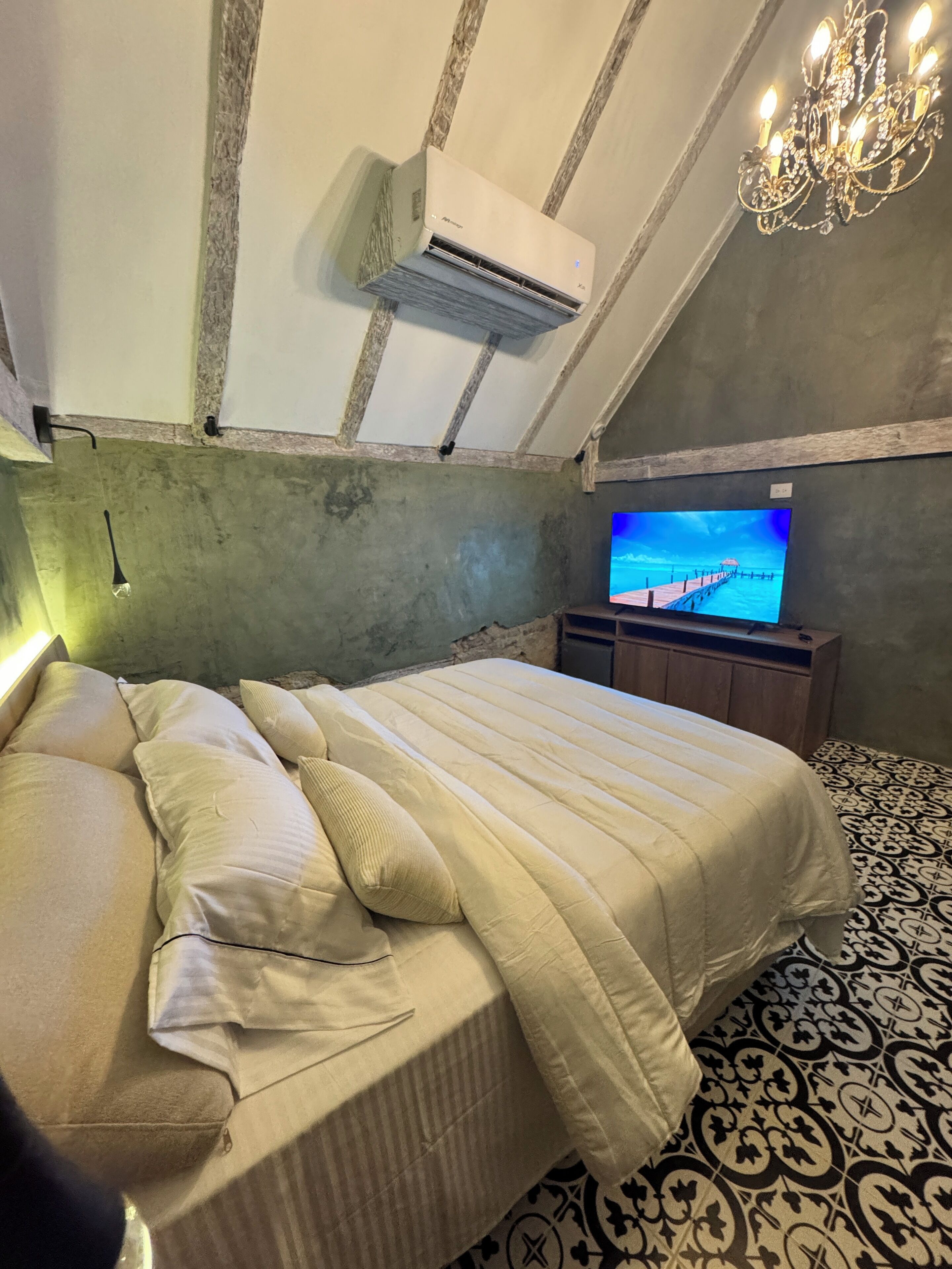 Un dormitorio con cama, televisión, aire acondicionado, una araña y suelo estampado.