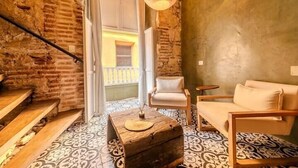 Un interior rústico con paredes de ladrillo visto, una escalera de madera, baldosas de suelo estampadas, un cofre de madera, un sillón blanco y una lámpara colgante.
