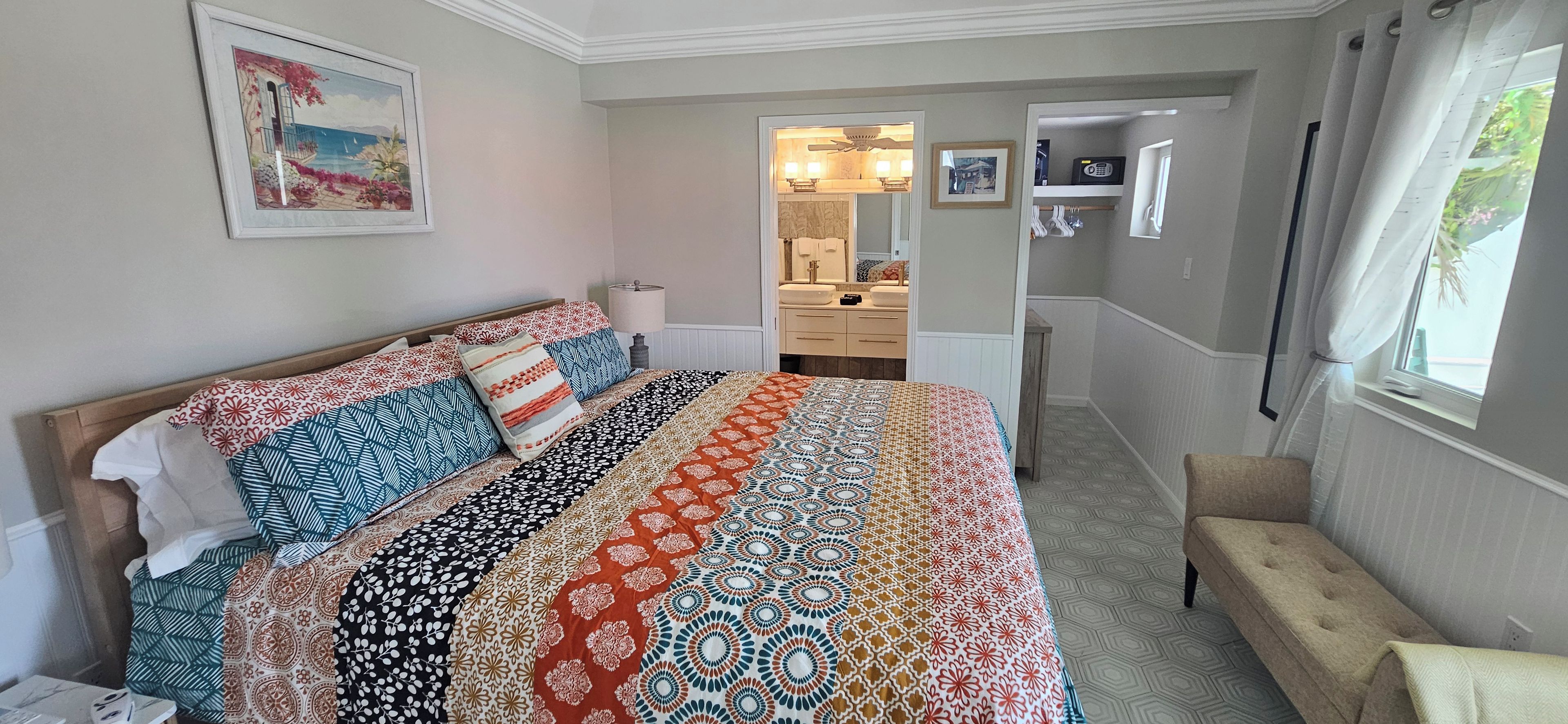 A bedroom with a large bed, a bedside table, a lamp, a chair, a window with curtains, a bathroom with a mirror, and a hallway leading to another room.