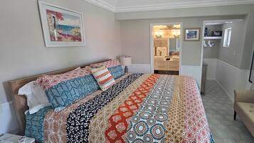 A bedroom with a large bed, a bedside table, a lamp, a chair, a window with curtains, a bathroom with a mirror, and a hallway leading to another room.