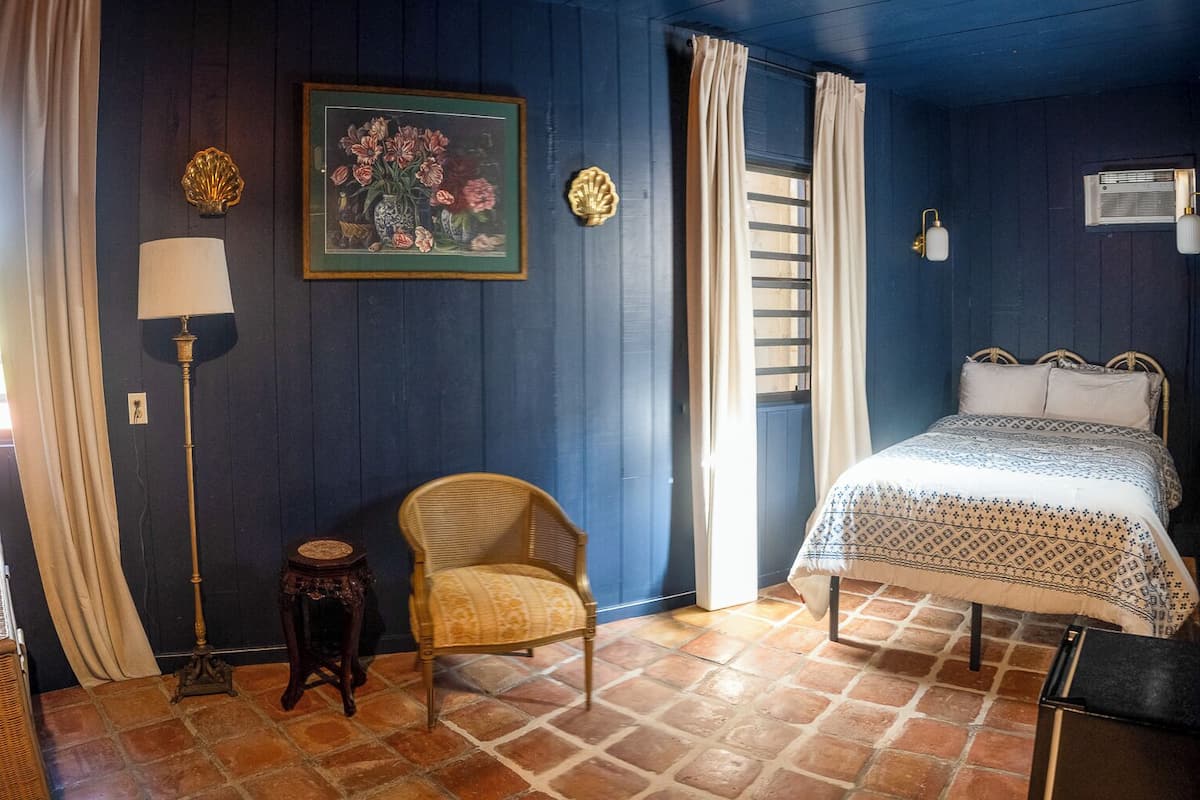 Una habitación con cama, una silla, una lámpara, un cuadro en la pared y una ventana con cortinas.