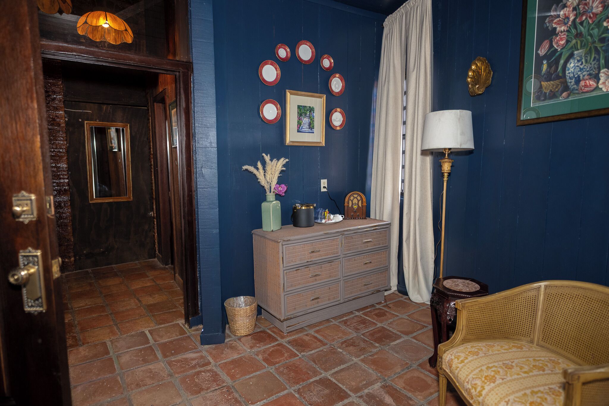 Een kamer met een houten deur, een dressoir, een lamp en een rieten stoel.