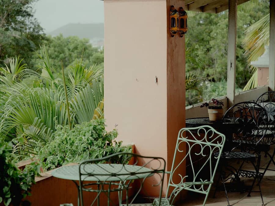 A covered patio area with metal chairs and tables, surrounded by lush greenery and a view of distant hills.