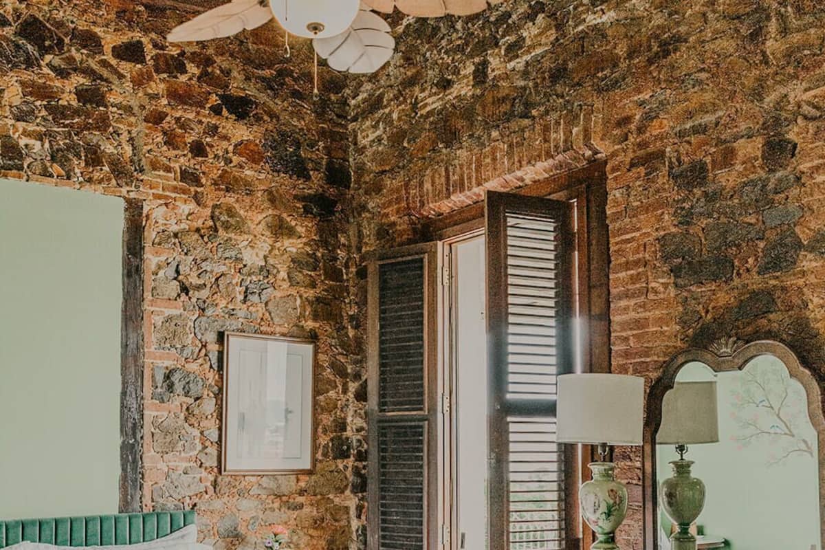 Un dormitorio con pared de piedra, una cama con cabecera verde, un aparador de madera, un ventilador de techo y una ventana con contraventanas.