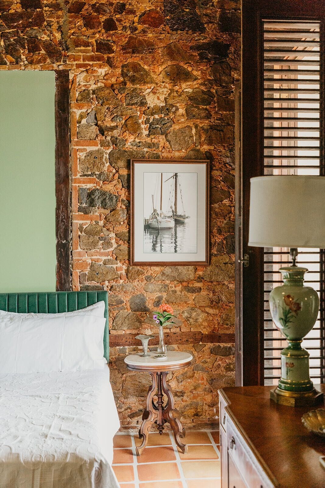 Habitación con pared de piedra, un cuadro enmarco de un barco, una mesita redonda con un florero, una cama con cabecera verde y una lámpara sobre una mesita de madera.