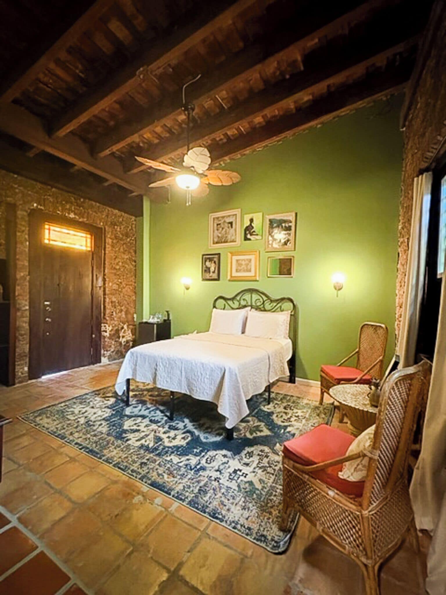 A room with a bed, a chair, a wall with framed pictures, a ceiling fan, and a door.