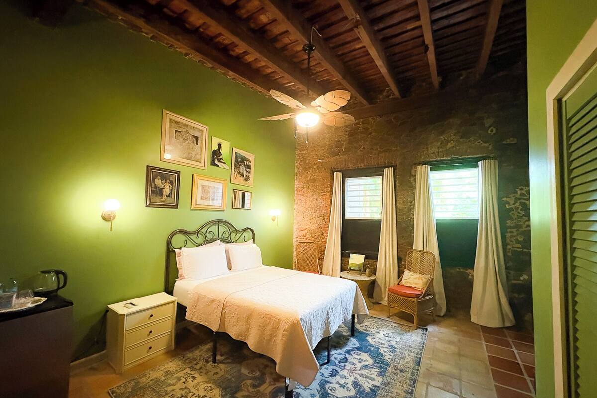 Un dormitorio con cama, mesitas de noche, ventilador de techo, ventana con cortinas e iluminación en la pared.