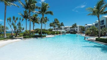 A large swimming pool surrounded by palm trees and a beach, with a building in the background.