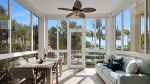A spacious sunroom with a ceiling fan, large windows, a dining area, and a view of a pool and palm trees. - Sunrise Retreat: Ocean & Pool Views, Gym, PB Courts, Game Room, Marina and more (Key Largo)