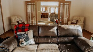 Een woonkamer met een bruine leren bank, een geruit plaid en een kussen met een ontwerp van een beer. Er staan twee stoelen met kussens, een houten deur en een uitzicht op een andere kamer met een eethoek.