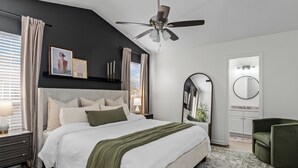 A modern bedroom with a large bed, a ceiling fan, a bedside table with a lamp, a green armchair, and a mirror on the wall.