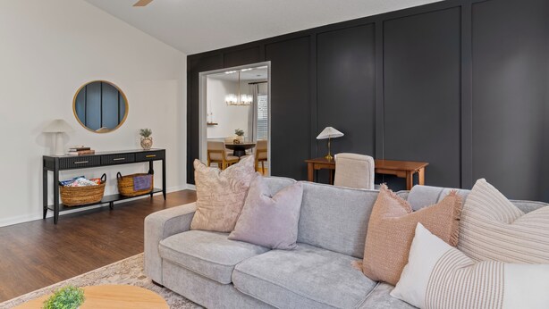 A living room with a grey sofa, a round mirror, a ceiling fan, and a dark wall.