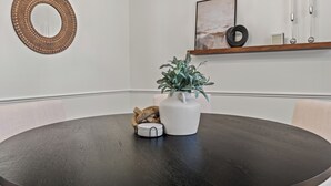A round dining table with a white vase and a small plant, a wooden side table with a basket, and a wall-mounted mirror.