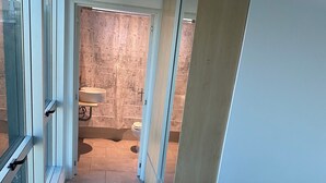 A narrow hallway with a bed on the right, a bathroom with a toilet and sink visible through a glass door, and a tiled floor.