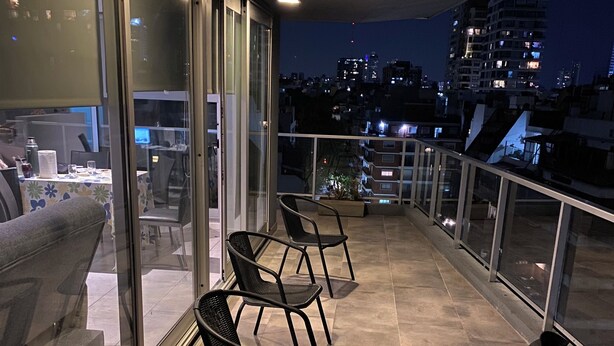 A balcony with four black metal chairs, a dining table with chairs, and a view of the cityscape at night.