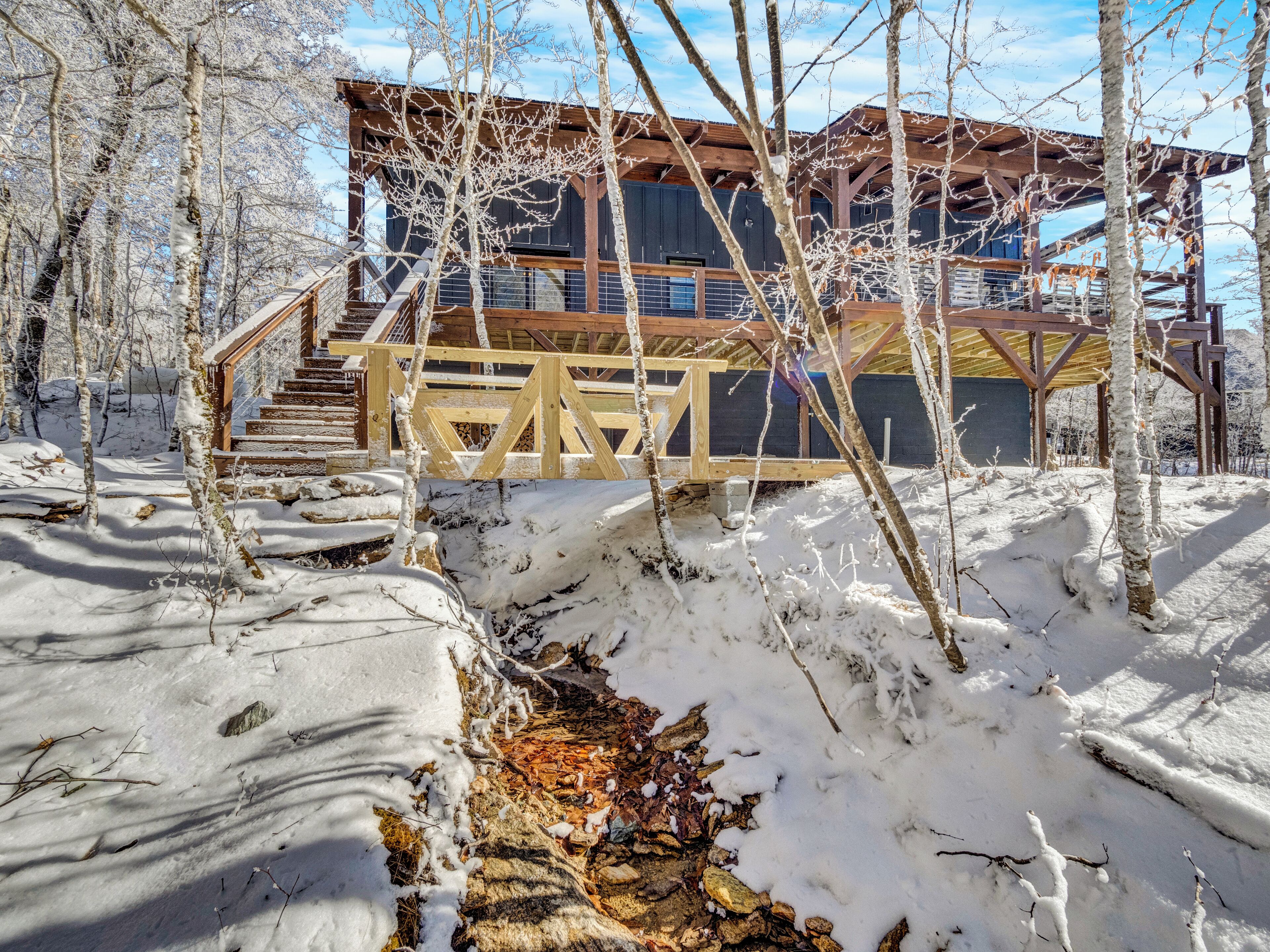 A wooden cabin with a staircase leading to a deck, surrounded by snow-covered ground and trees.
