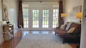 Un salón amplio con piso de madera, un sofá de cuero marrón, sillas de plástico transparente, un tapete blanco y un gran ventanal que da al exterior.