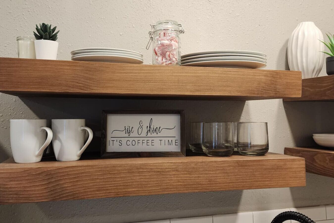 Una estantería de madera con tazas blancas, un tarro de cristal y un letrero que dice "rise & shine, it's coffee time".