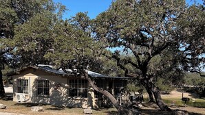 A rustic cabin with a large tree in front, surrounded by a gravel driveway and a grassy area. - Riverbend on the Frio Cabin 4 (Concan)