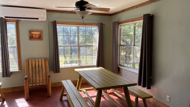 Ein Zimmer mit Holztisch und Bänken, großen Fenstern, einem Deckenventilator und einer Klimaanlage.