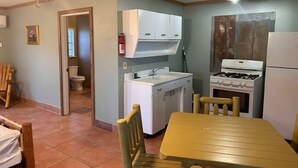 Cocina compacta con armarios blancos, estufa y lavaplatos. Cuenta con una mesa de madera y sillas.