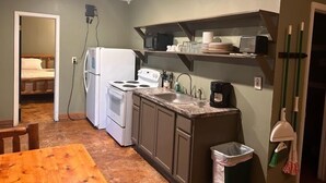 Eine kompakte Küche mit Kühlschrank, Herd, Mikrowelle und Spüle. Ein Holztisch und ein Mülleimer sind ebenfalls vorhanden.